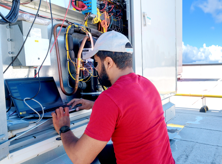 Worker on rooftop working on laptop with an open electrical panel revealing wires